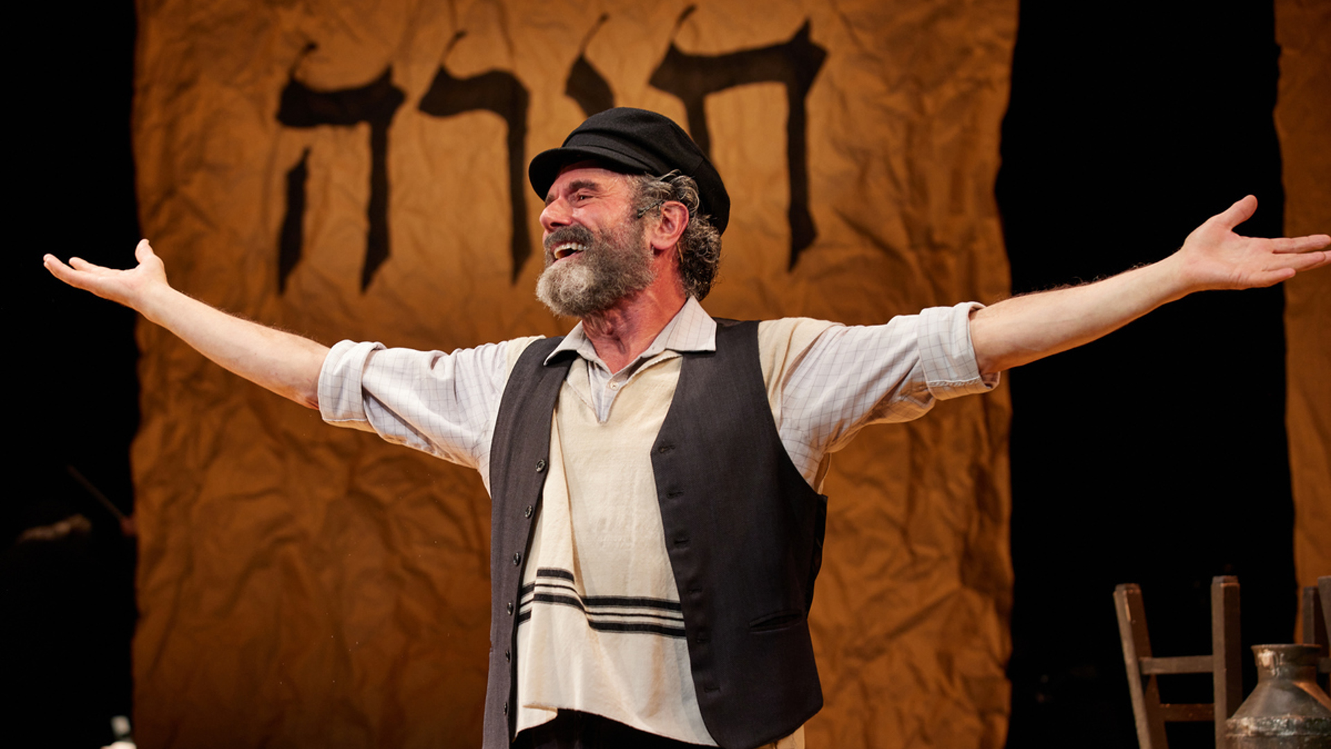 A man stands on stage stands with his arms out, he is smiling at the audience. He is wearing a black hat and a gray vest as well as a tallit.
