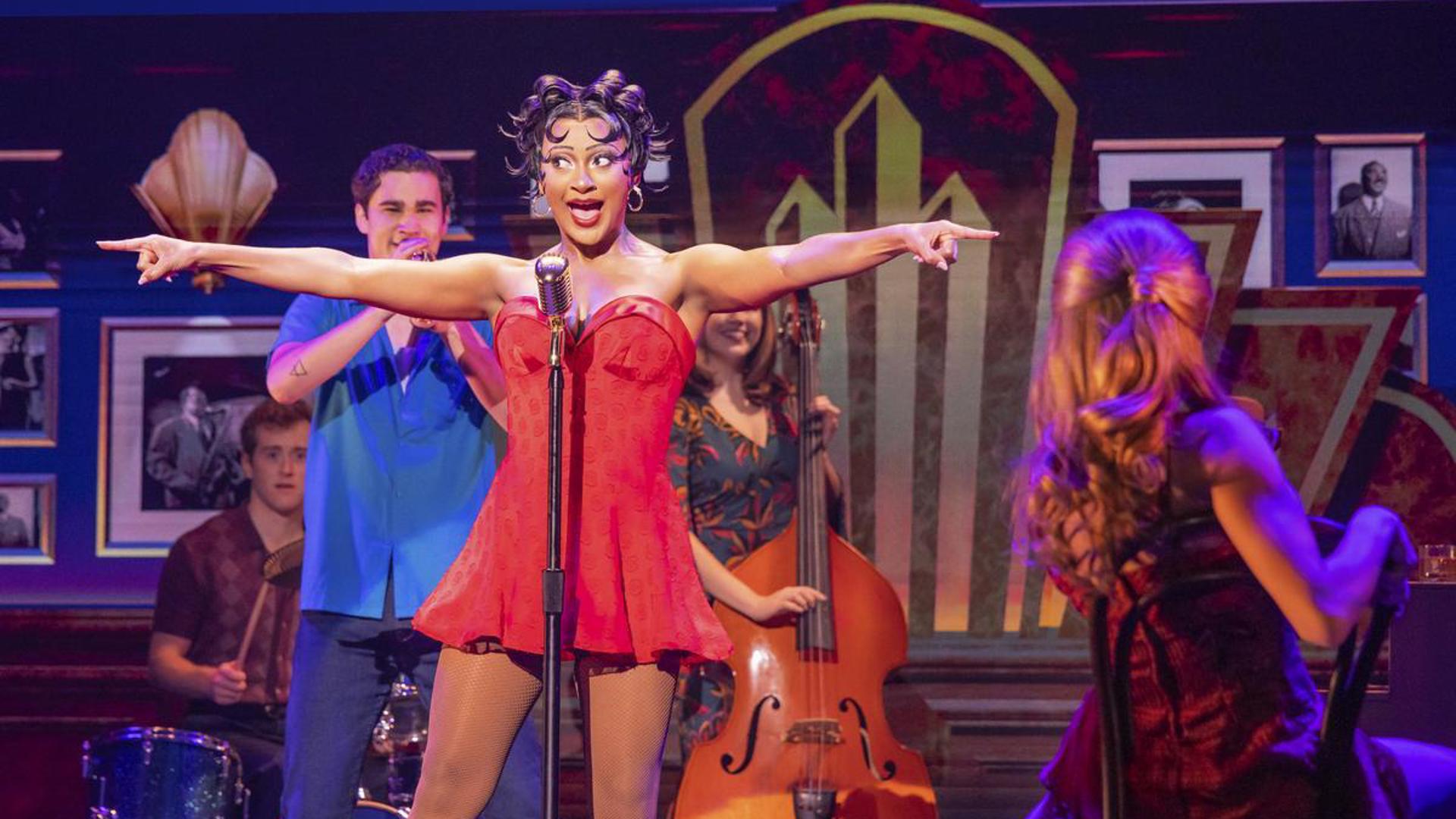 An actress stands at a microphone, looking at the audience and singing. She is wearing a short, red dress and has her arms pointed out to either side. There are actors playing instruments behind her and an actor sitting on chair, looking at her.