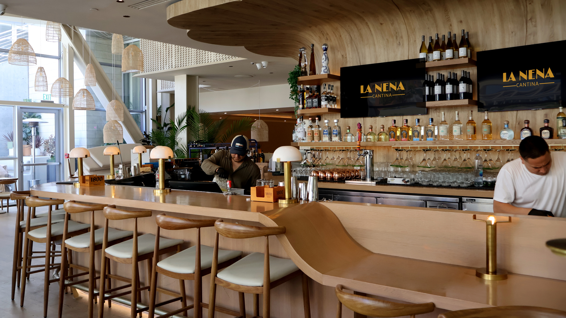 Interior of La Nena. A view of the bar with wooden countertops and chairs. Two workers are seen making drinks behind the bar.