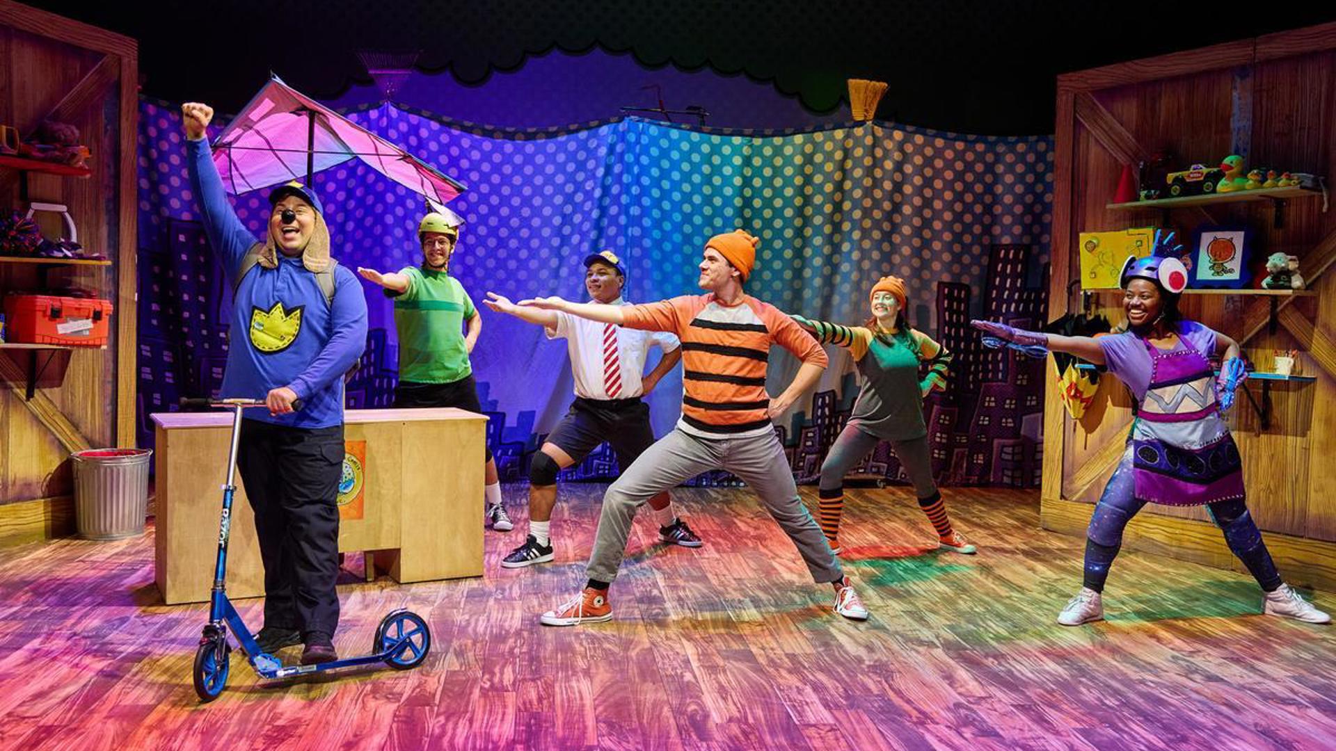 An actor stands on stage dressed as dog man, they are on a blue scooter, smiling with one fist raised in the air. Five other actors stand scattered around them, lunging toward dog man and smiling with their right hand extended and their left on their hip.