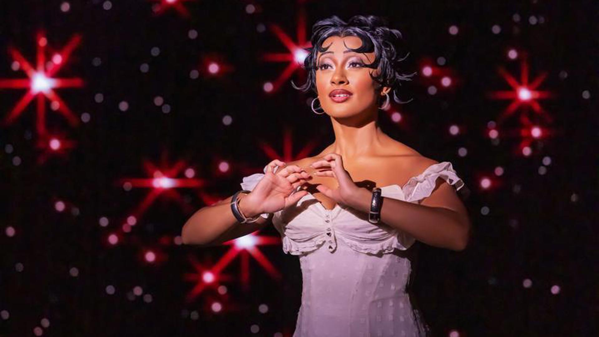 An actress stands on stage with both arms held in front of her, looking out to the audience. She is wearing a knee length, sleeveless white dress and behind her there is a starry sky.
