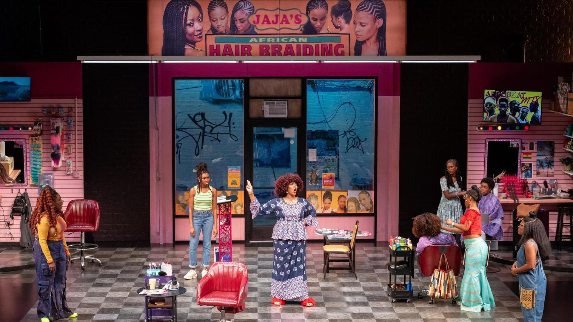 Eight performers stand onstage. One is center, speaking animatedly while others watch.