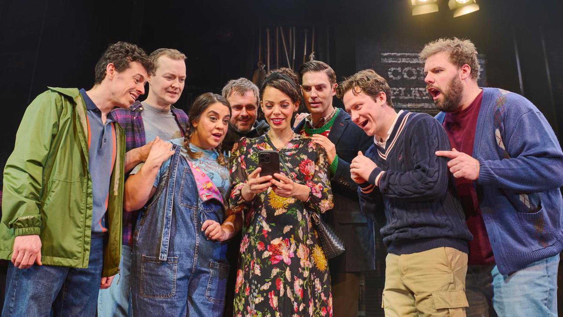 Eight performers stand huddled together, looking at a phone.