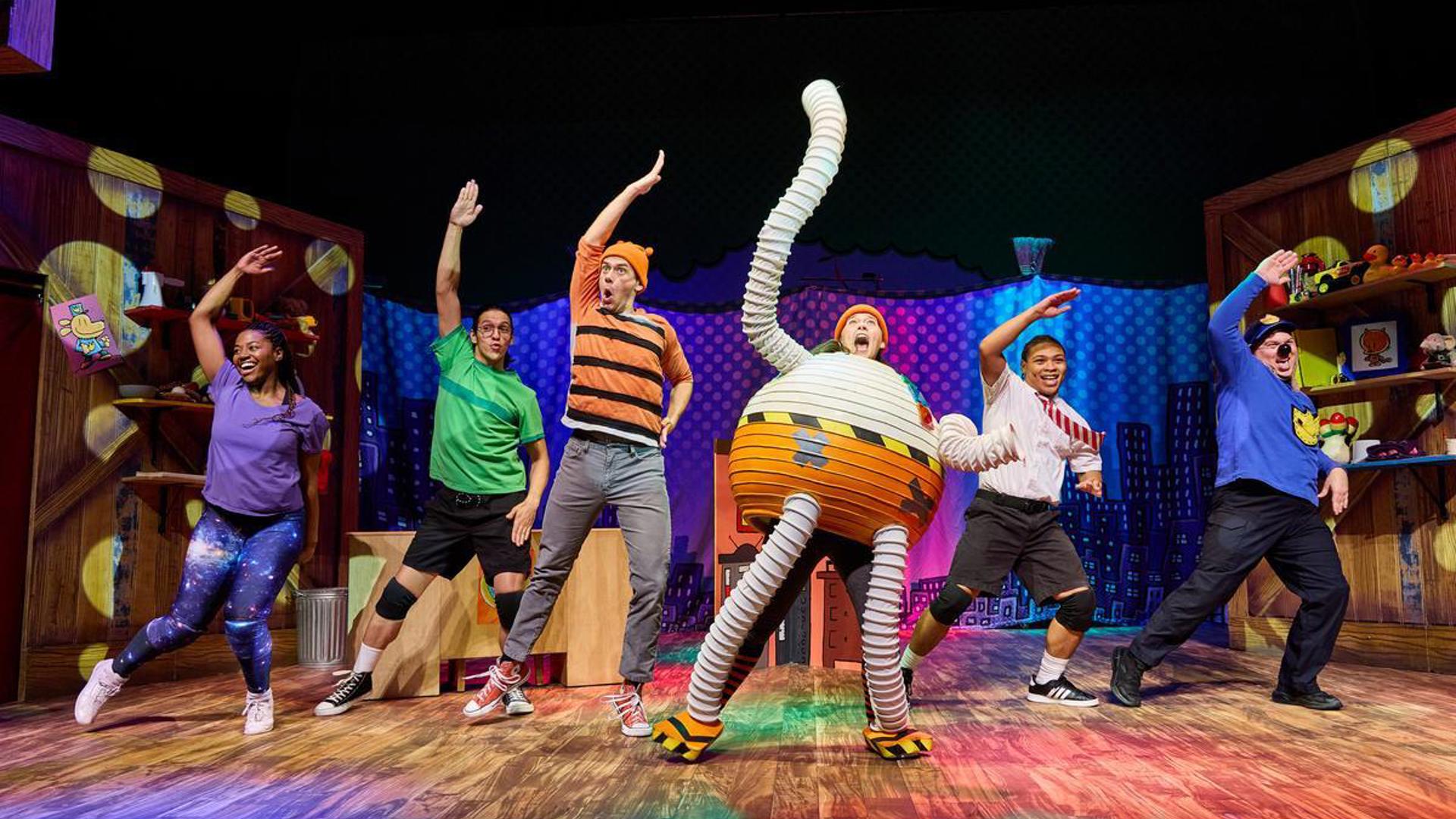 There are six actors on stage dancing in a line, all of them are extending their right arms to the sky over their heads.