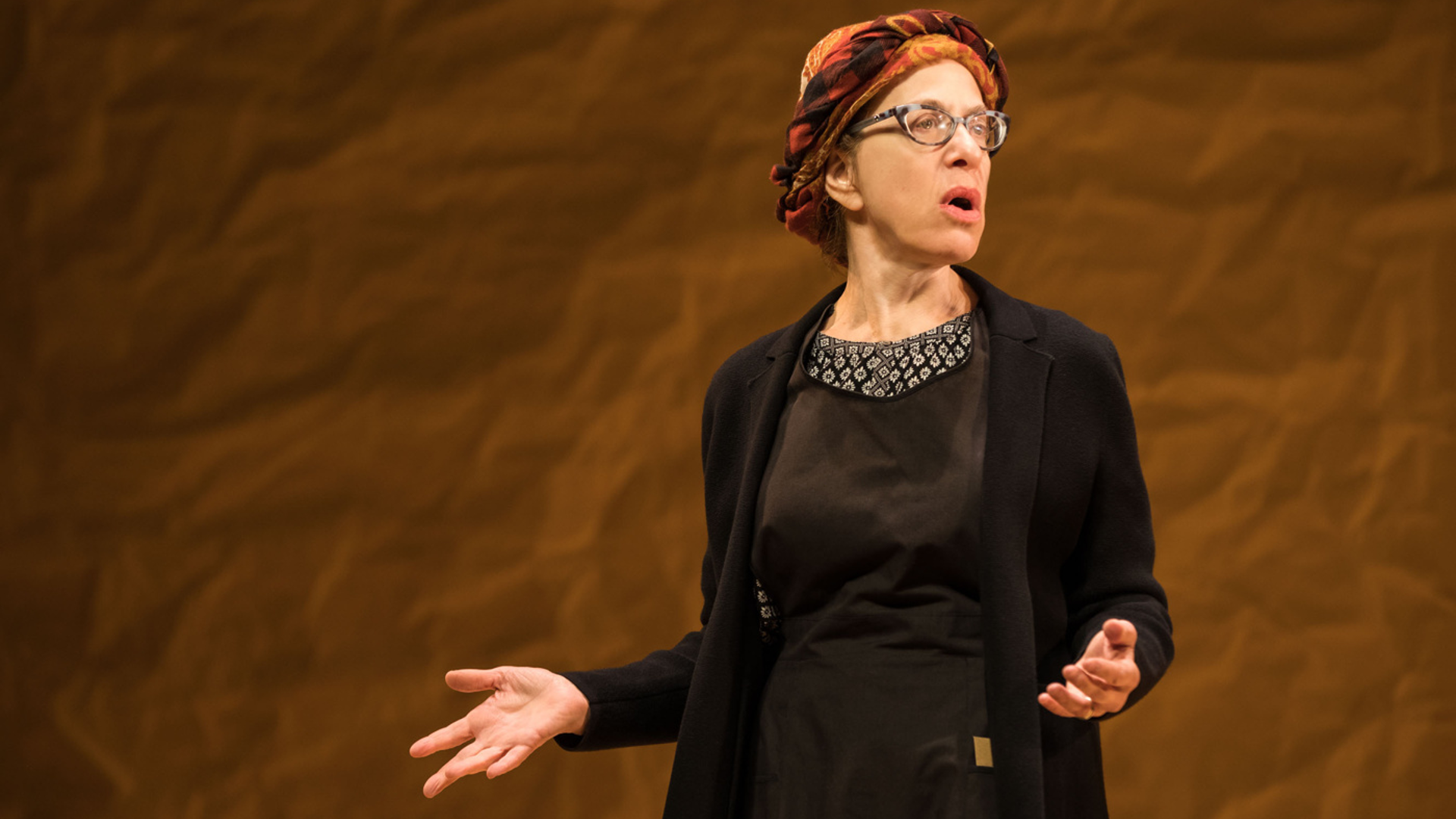 A woman on stage stands with both arms extended to the audience looking to the side.