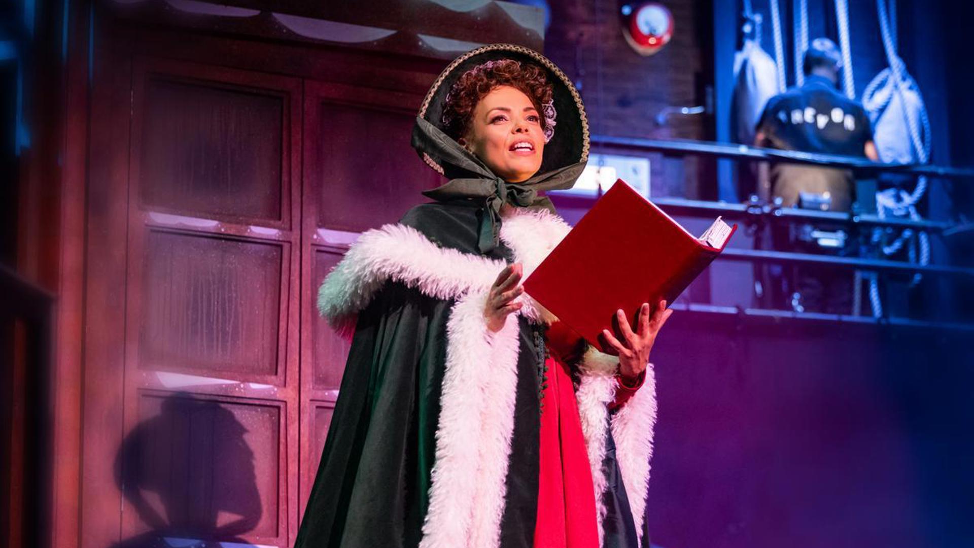 A performer stands on stage in a fur cloak, reading a book aloud.