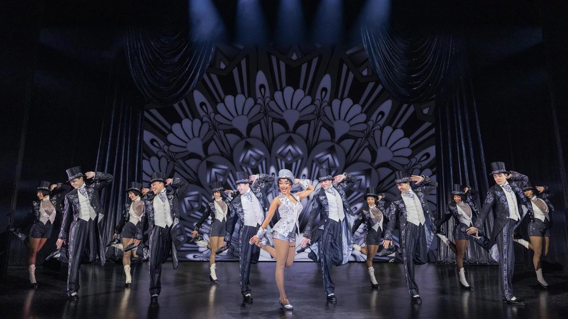 A company of actors all dance in top hats and suits. They have their right  hand on their left heel and their left hand on their head. They look towards their right side.
