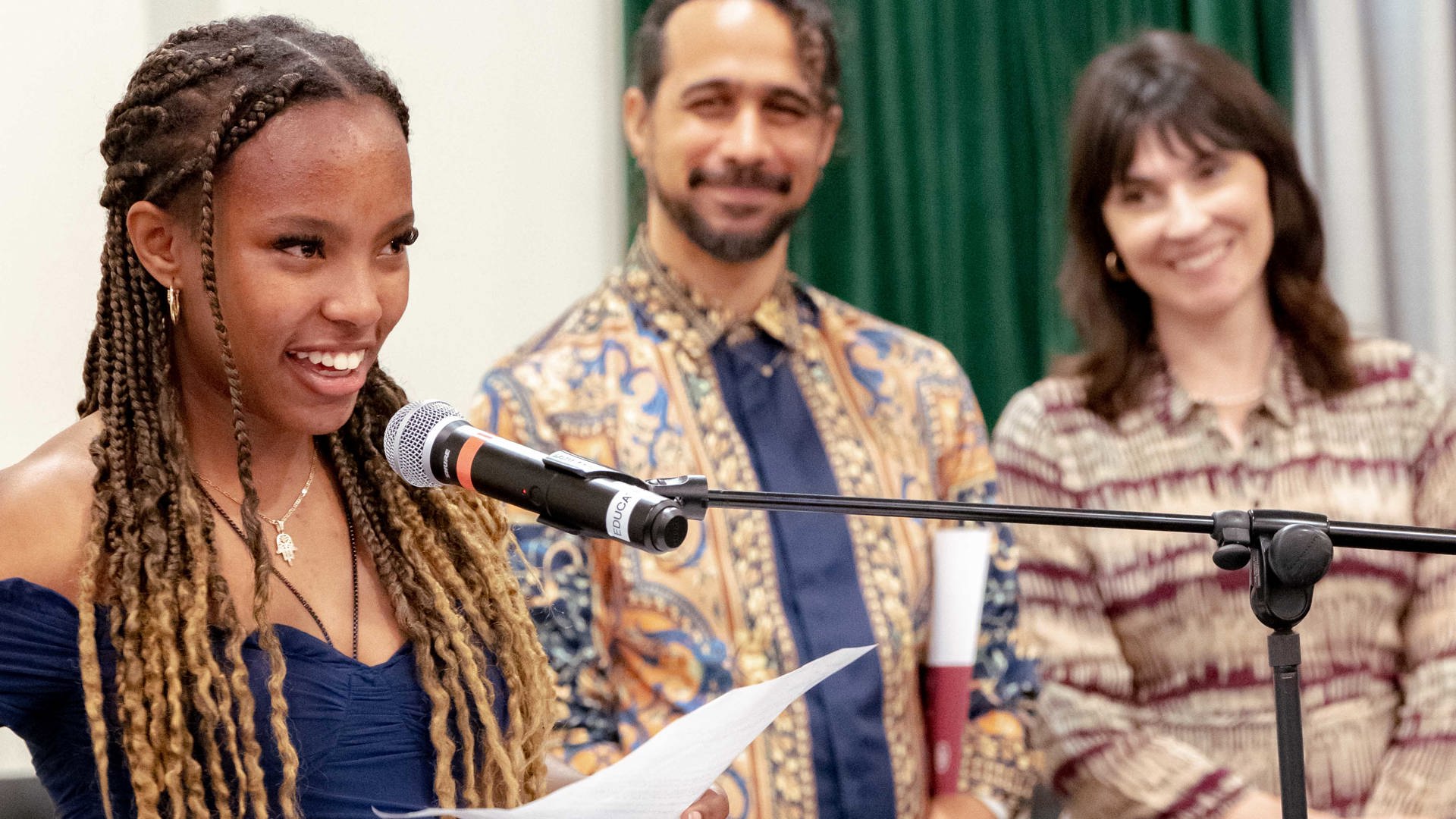 A person delivering a speech into a microphone, two other people stand in the background smiling. 