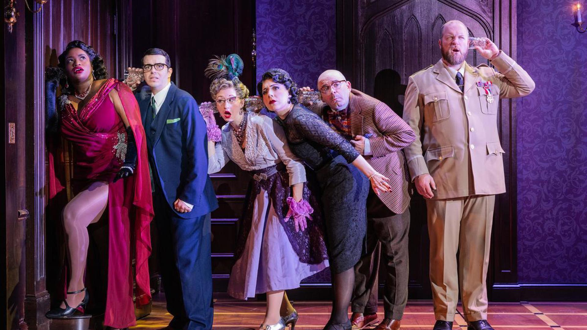 Six actors stand on stage in a line on one side of a closed wooden door, five of them are crouched close together with their ears pressed against glasses, trying to hear a conversation inside the closed door. The other actor is standing facing away from the rest, his glass pressed to his own ear, but the other end is connected to nothing.
