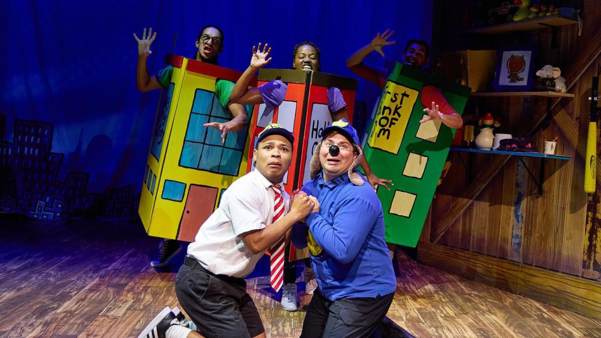 Two actors kneel on stage, holding each others hands and looking out at the audience. Three actors stand behind them in a line, dressed as buildings and reaching for them.