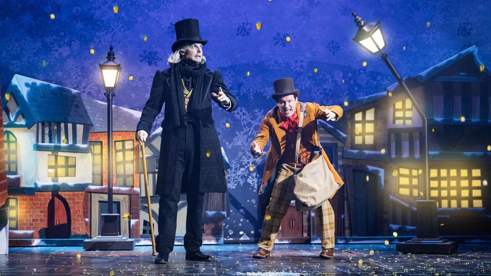 Two performers stand on a snowy straight in front of a row of houses.