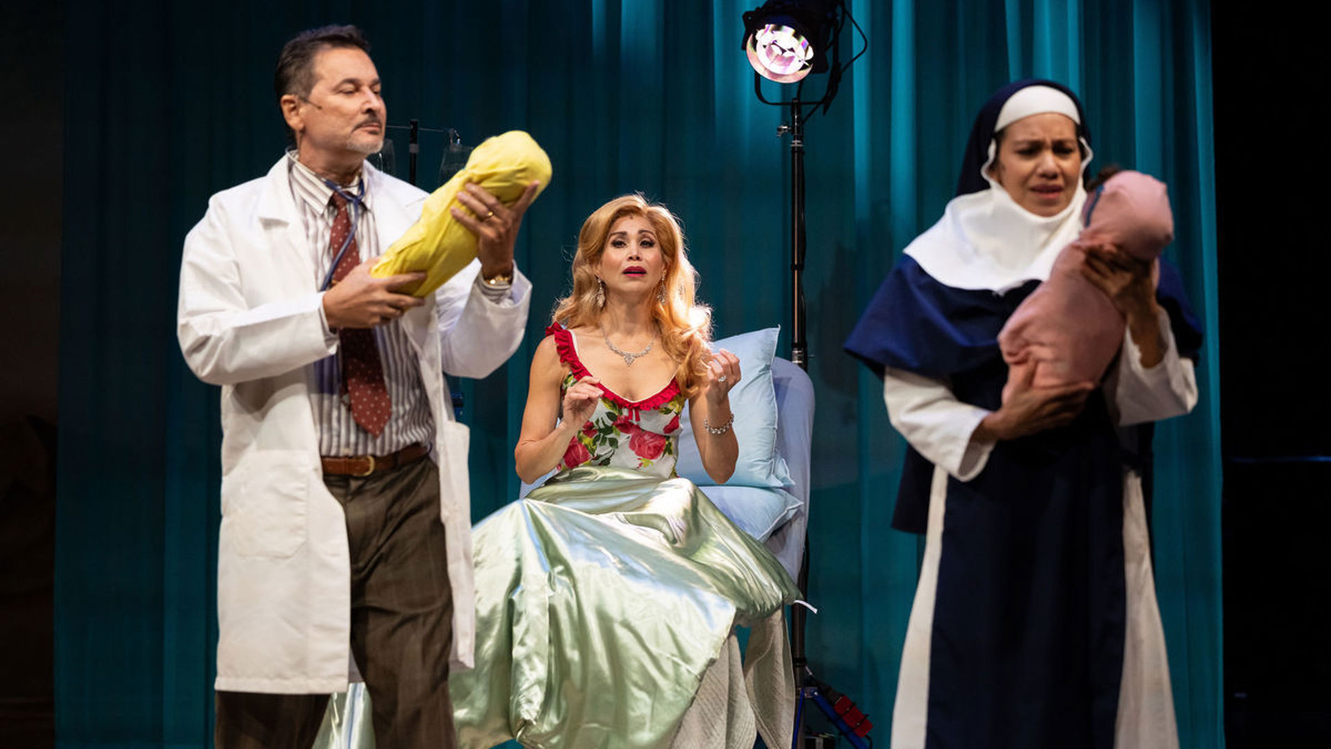 An actor dressed as a doctor holds a bundle that resembles a newborn baby. An actor dressed as a nun holds a similar bundle. Between them, an actor watches from a hospital bed looking distressed.