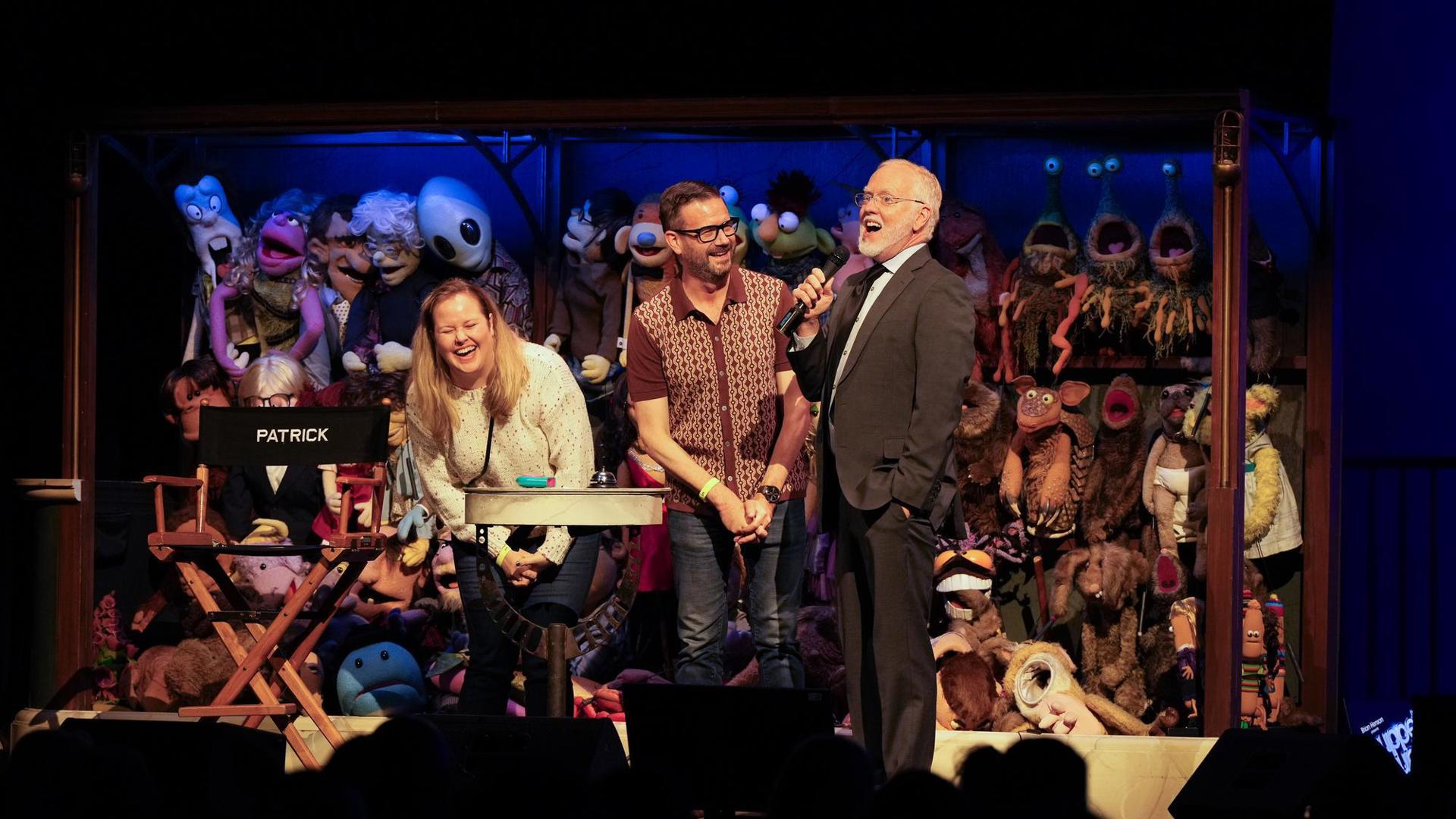 actors laughing on stage in front of a wall of puppets