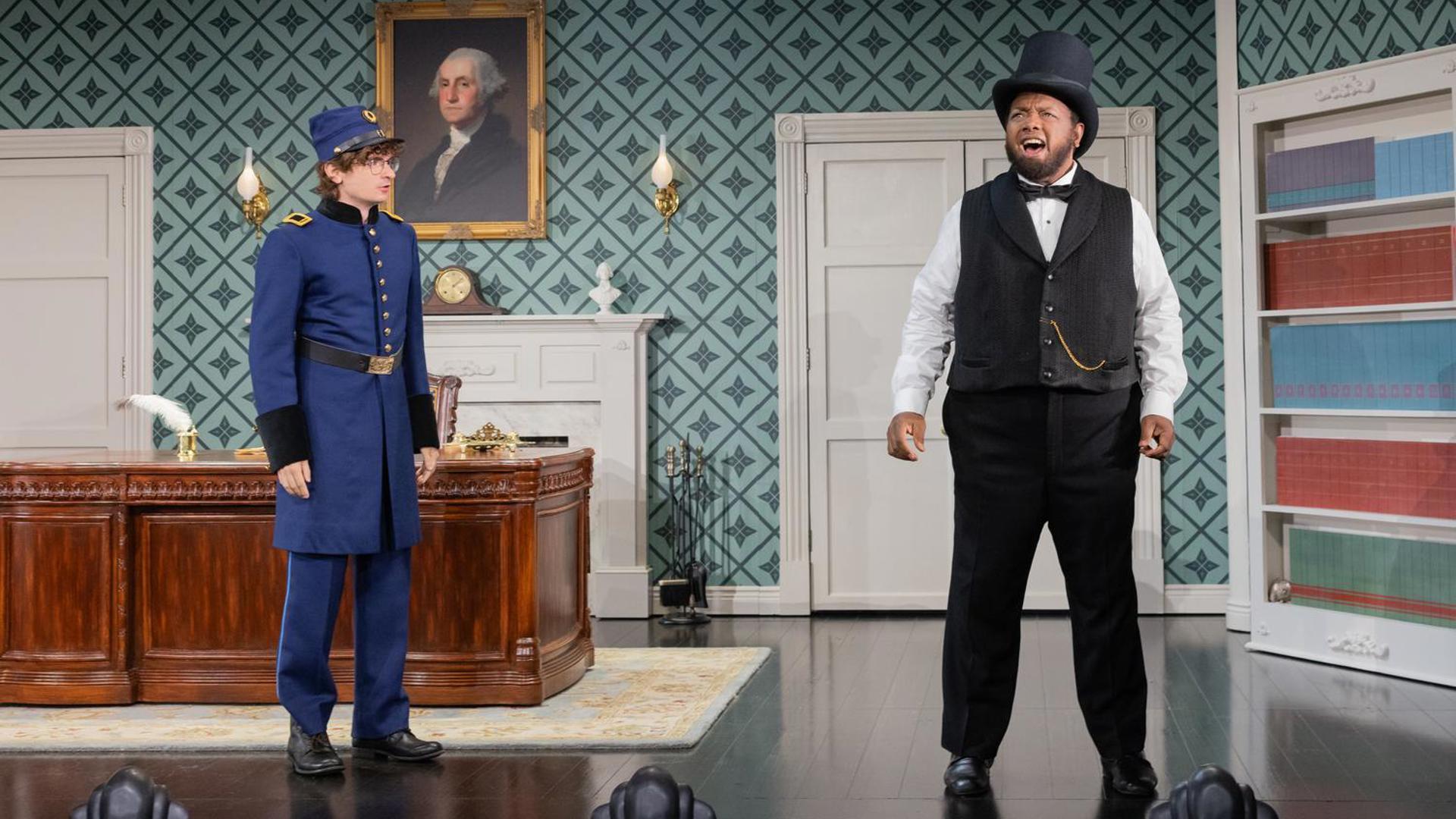 An actor stands in a black top hat, speaking to the audience. To his right, another actor in a blue soldier's outfit looks at him.