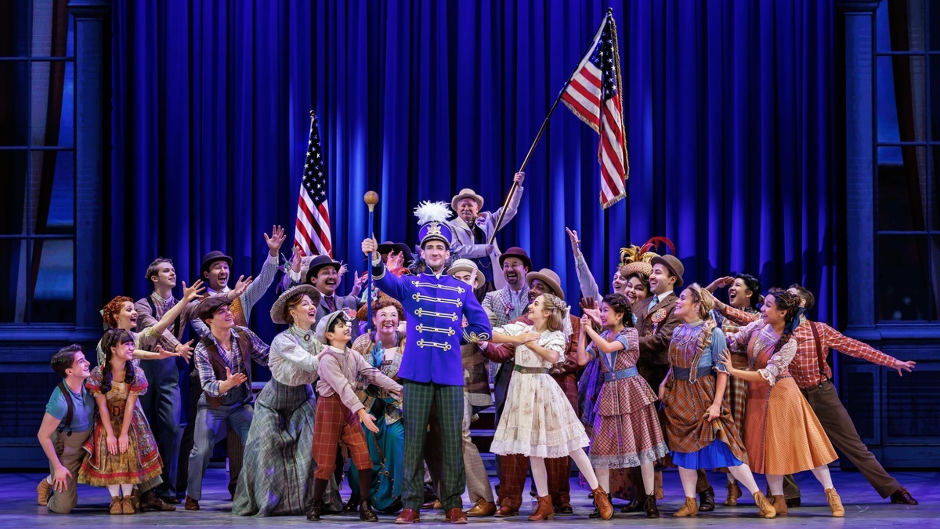 A man stands in the center of the stage dressed as a band conductor, raising a baton. A crowd of people is gathered around him, leaning in. One actor in the back is raising an American flag.