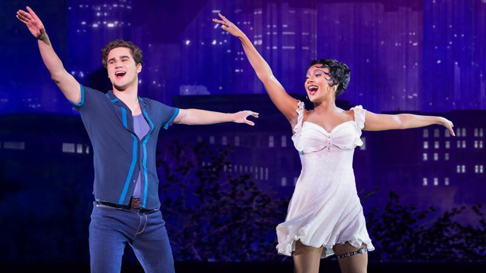 Two actors dance on stage in front of a cityscape at night. They both have their right arm extended above and in front of them and their left arm extended behind them, looking towards their right hands.