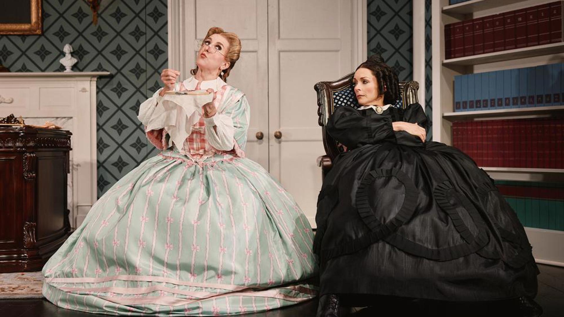 Two performers sitting side by side in chairs, one is sewing and the other is watching her.