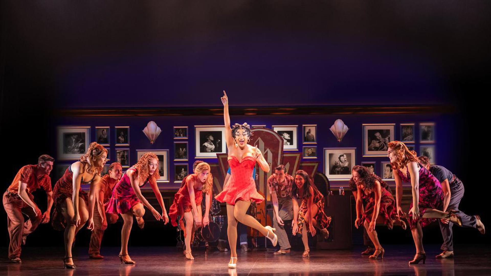An actress dances on stage in a short, red dress. She has her right hand above her head pointing at the sky and her left heel lifted off the ground. She is looking out at the audience and singing while company members dance behind her.