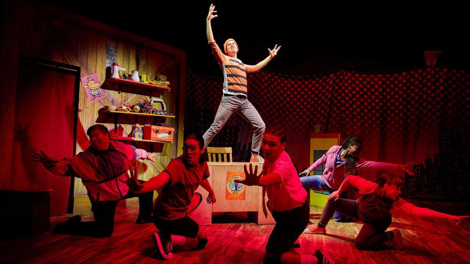 An actor stands on a desk under a spotlight, arms extended toward the sky. Five actors surround them in a semi-circle under red light, reaching away from the actor on the desk.