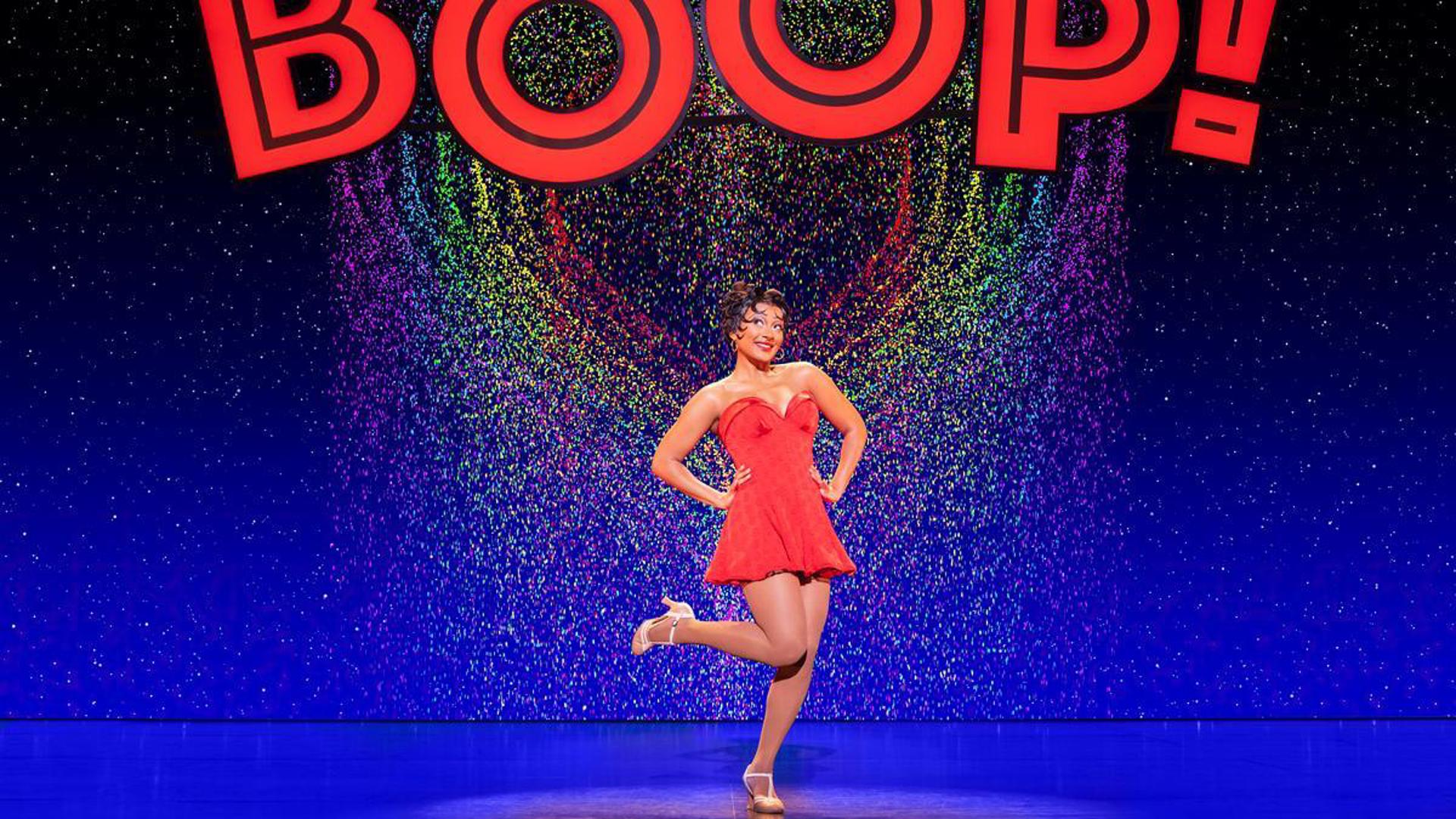 An actress stands on stage in a short, red dress. She has her hands on her hips and one heel lifted to her right. She is looking up to her right. Above her is a rainbow heart and the word 'BOOP!' in red text.