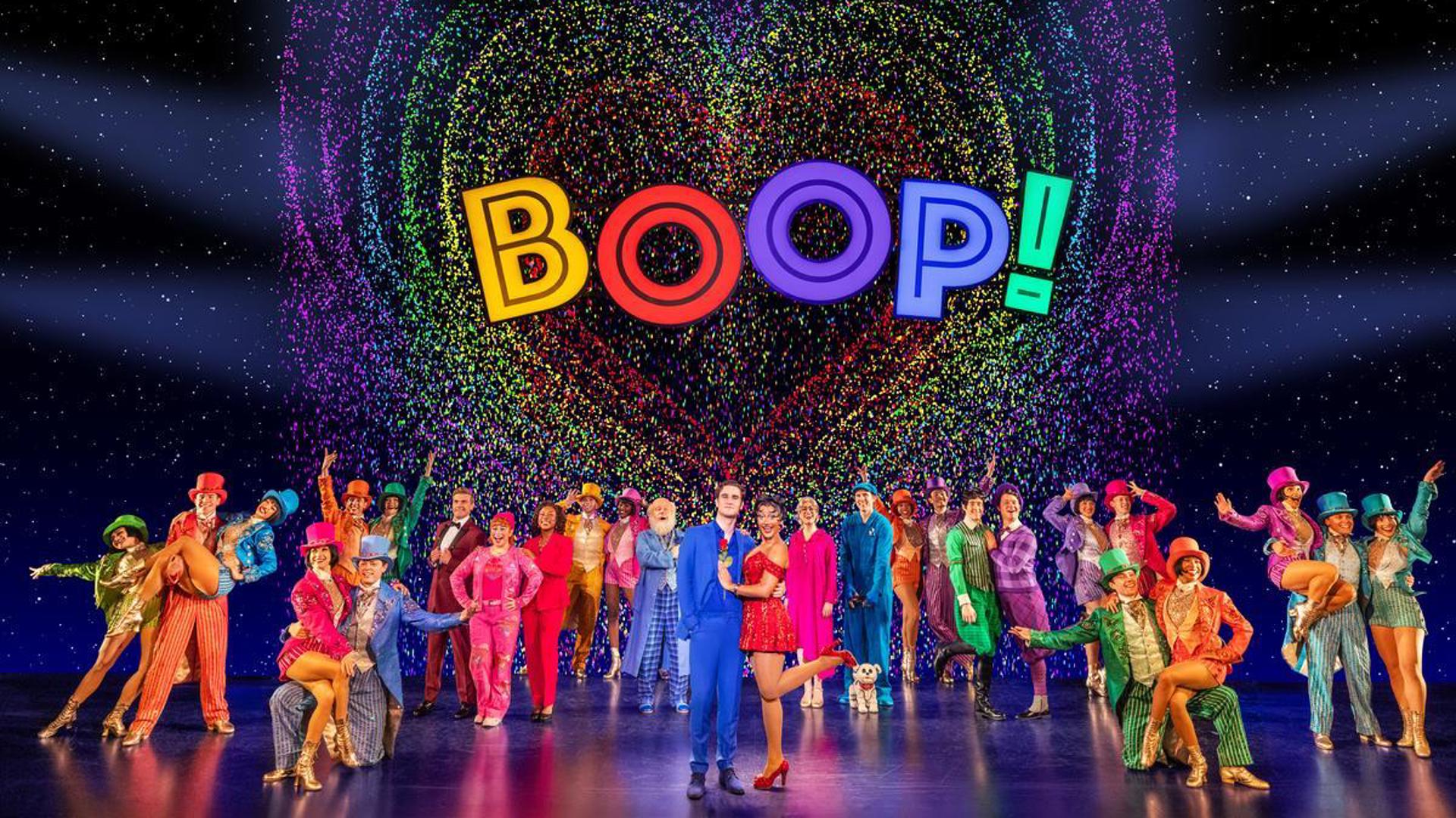 A cast of actors all stand, posing and looking out at the audience, dressed in colorful outfits. Behind them is the word 'BOOP!' in a rainbow heart.