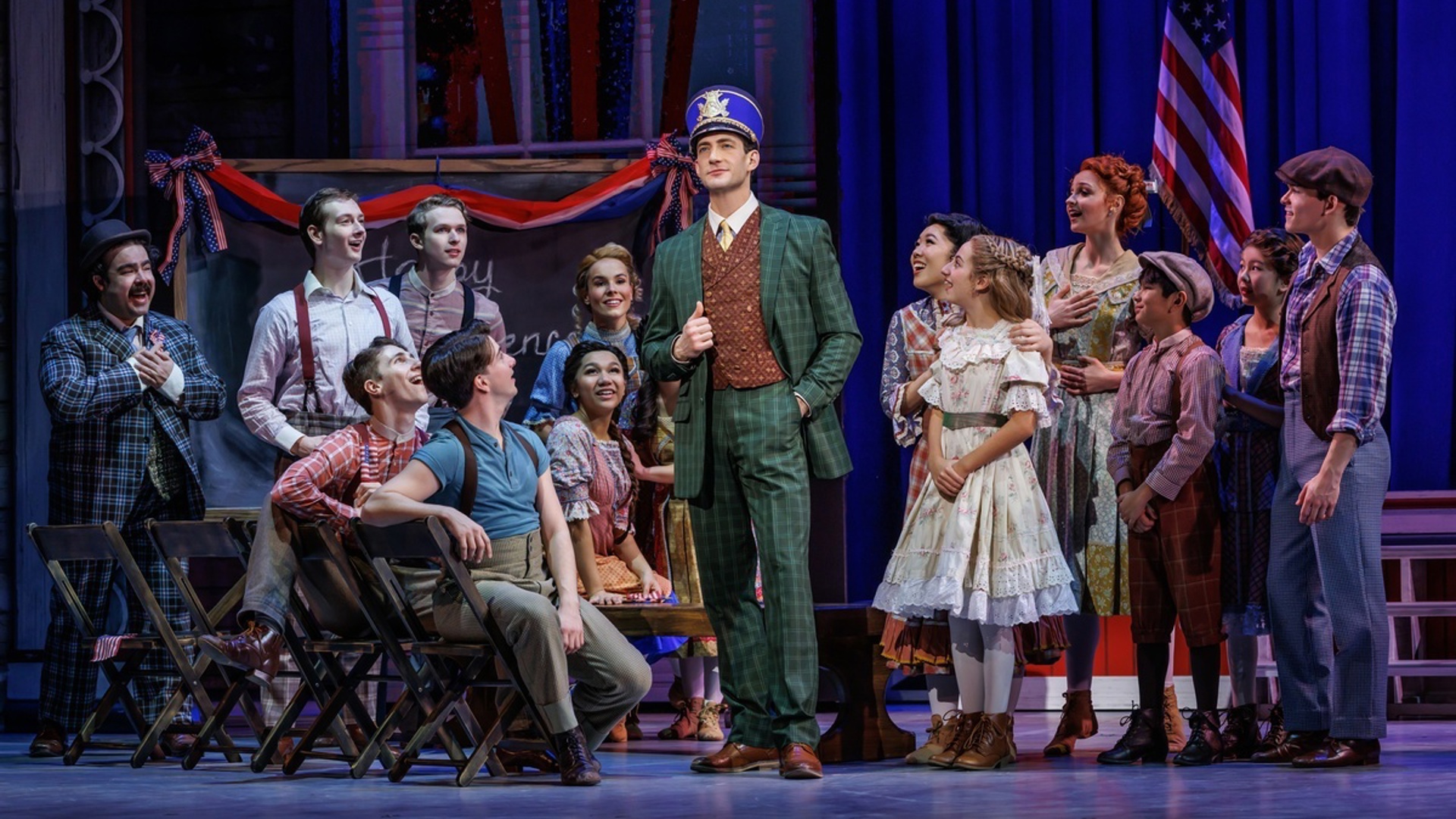 An actor stands in the center of a semicircle of other actors, looking out at the audience wearing a suit and a band cap (shako). The rest of the actors look in at him, smiling.
