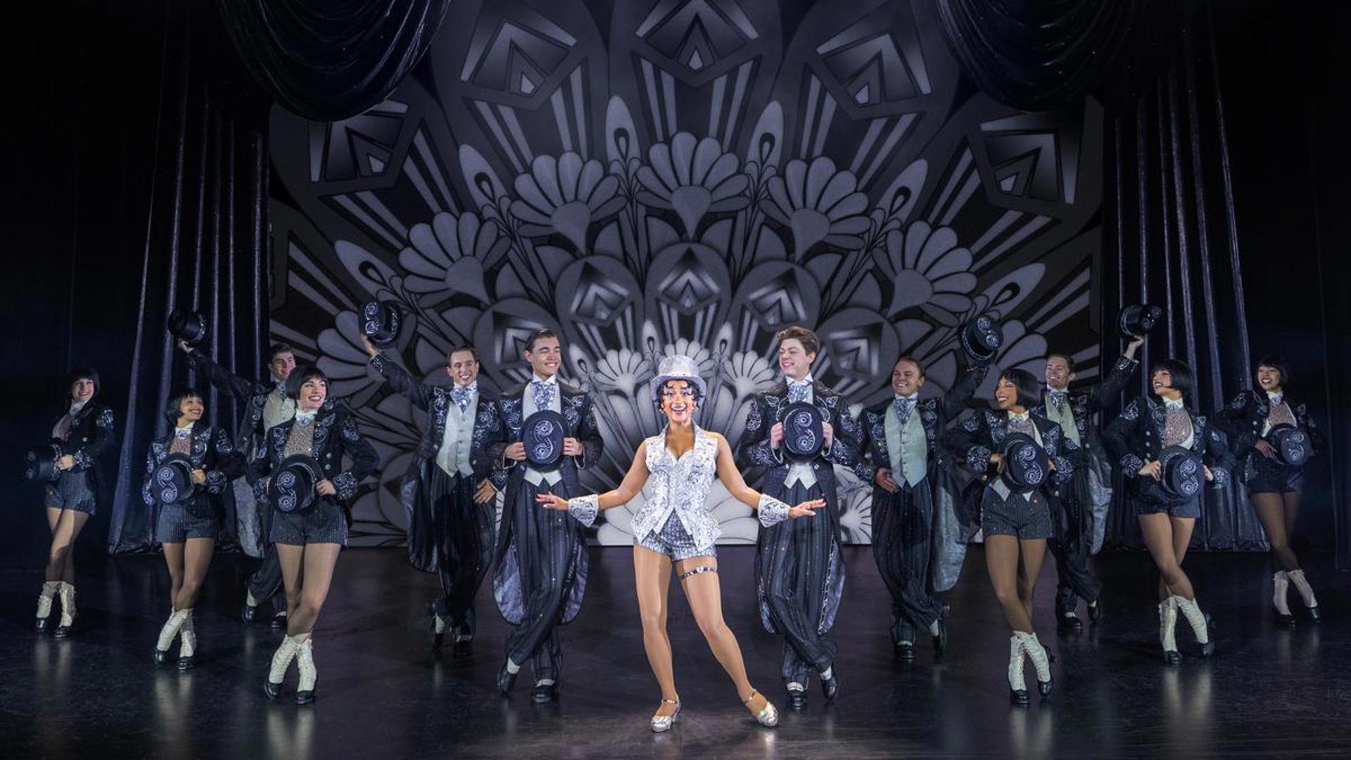 An actress stands at the center of the stage. A group of dancers are lined up around her, holding top hats and looking toward the actress in the center.