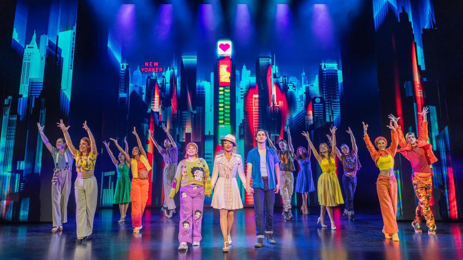 Three actors stand in the center of the stage, holding hands. A company of actors dance around them wearing multicolored outfits in front of a New York city scape.