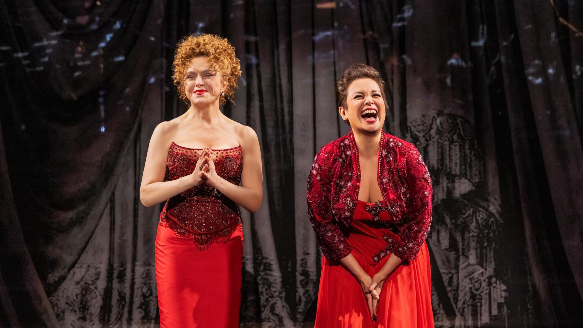 Two performers in red stand in front of a curtain. One clasps her hands together and the other is laughing.