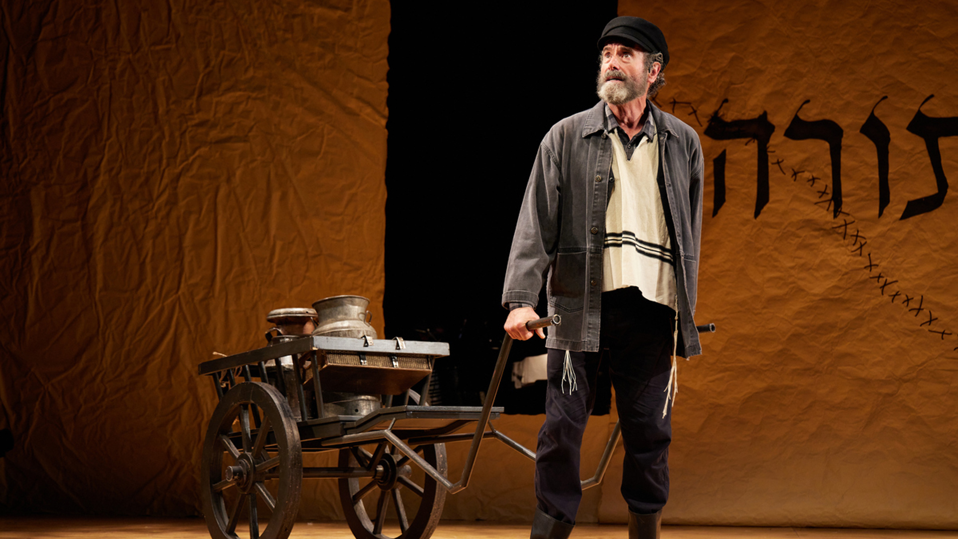 A man stands on stage carries a wagon behind him with two metal jugs inside. He is wearing a black hat and a gray vest as well as a tallit.