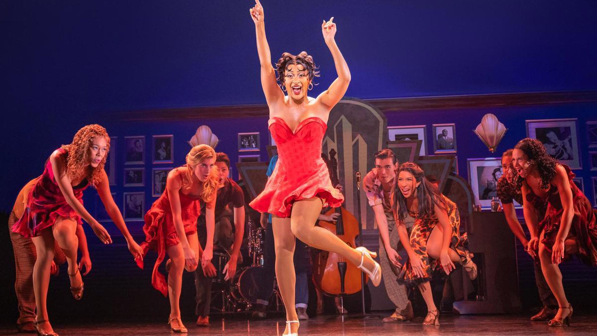 An actress dances on stage in a short, red dress. She has both her hands above her head pointing at the sky and her left heel lifted off the ground. She is looking out at the audience and singing while company members dance behind her.