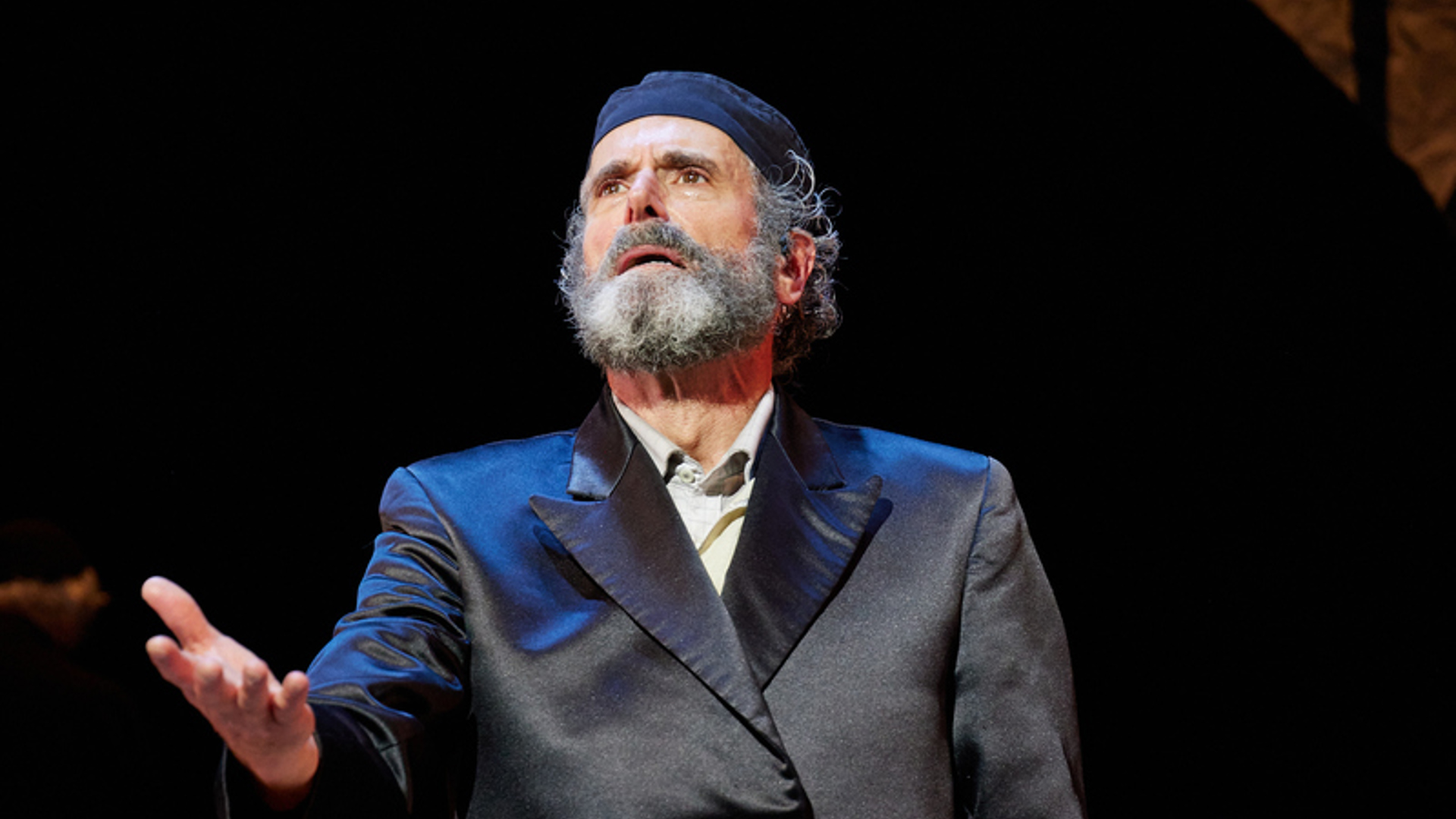 A man stands on stage stands with his right arm extended to the audience. He is wearing a black hat and a black trench coat. He is holding a brown book in his left hand.