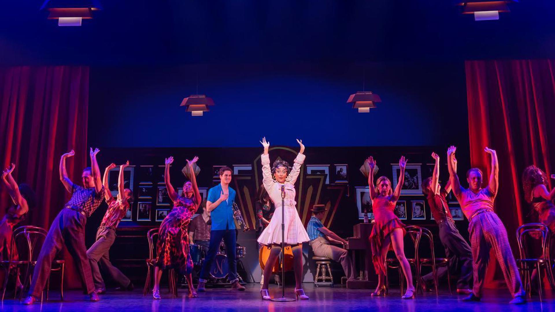 A company of actors dance on stage in a club. An actress stands at a microphone in the middle wearing a long sleeve white coat. To her right, an actor holding a trumpet has his hand on his heart, looking at her.