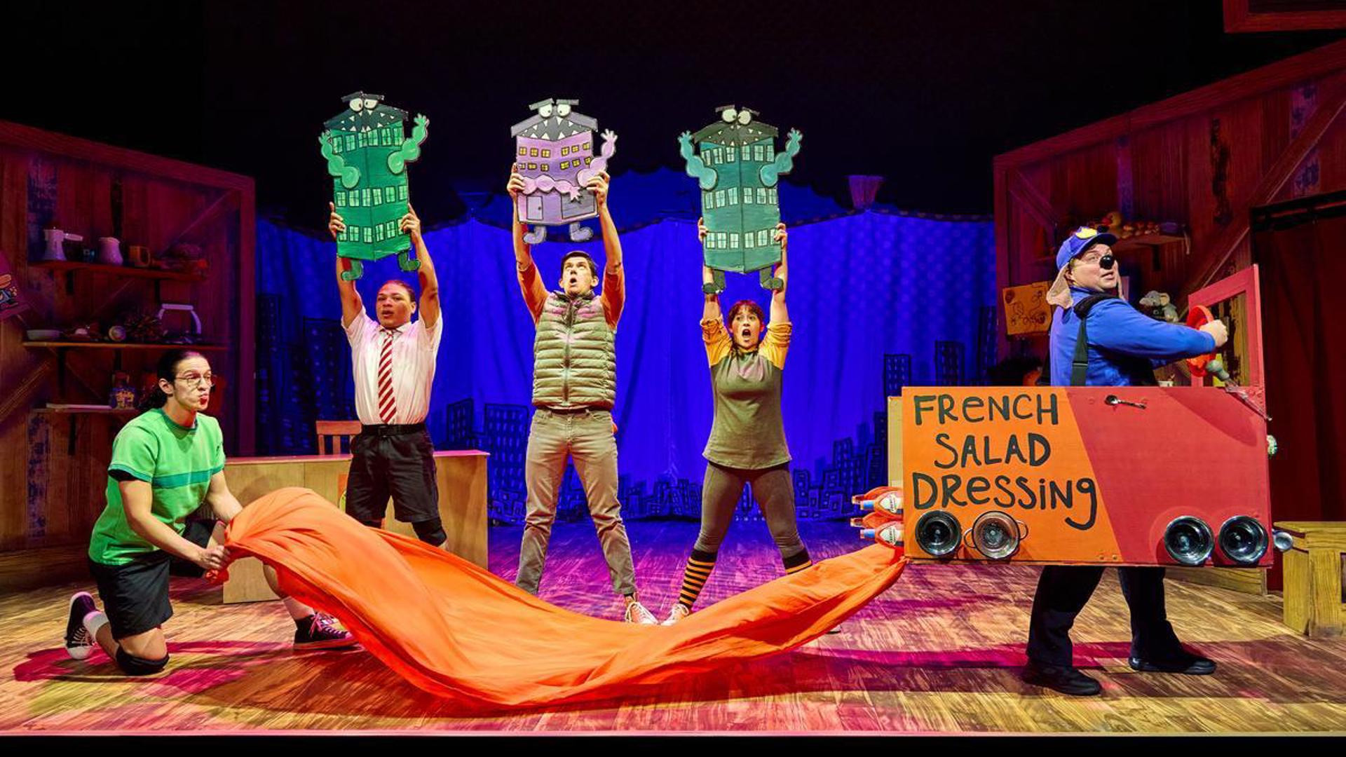 An actor drive a truck labeled "French Salad Dressing", another actor holds on to a long train of orange fabric behind the truck. Three actors stand behind them holding cartoon drawings of buildings with mouths and faces on them.