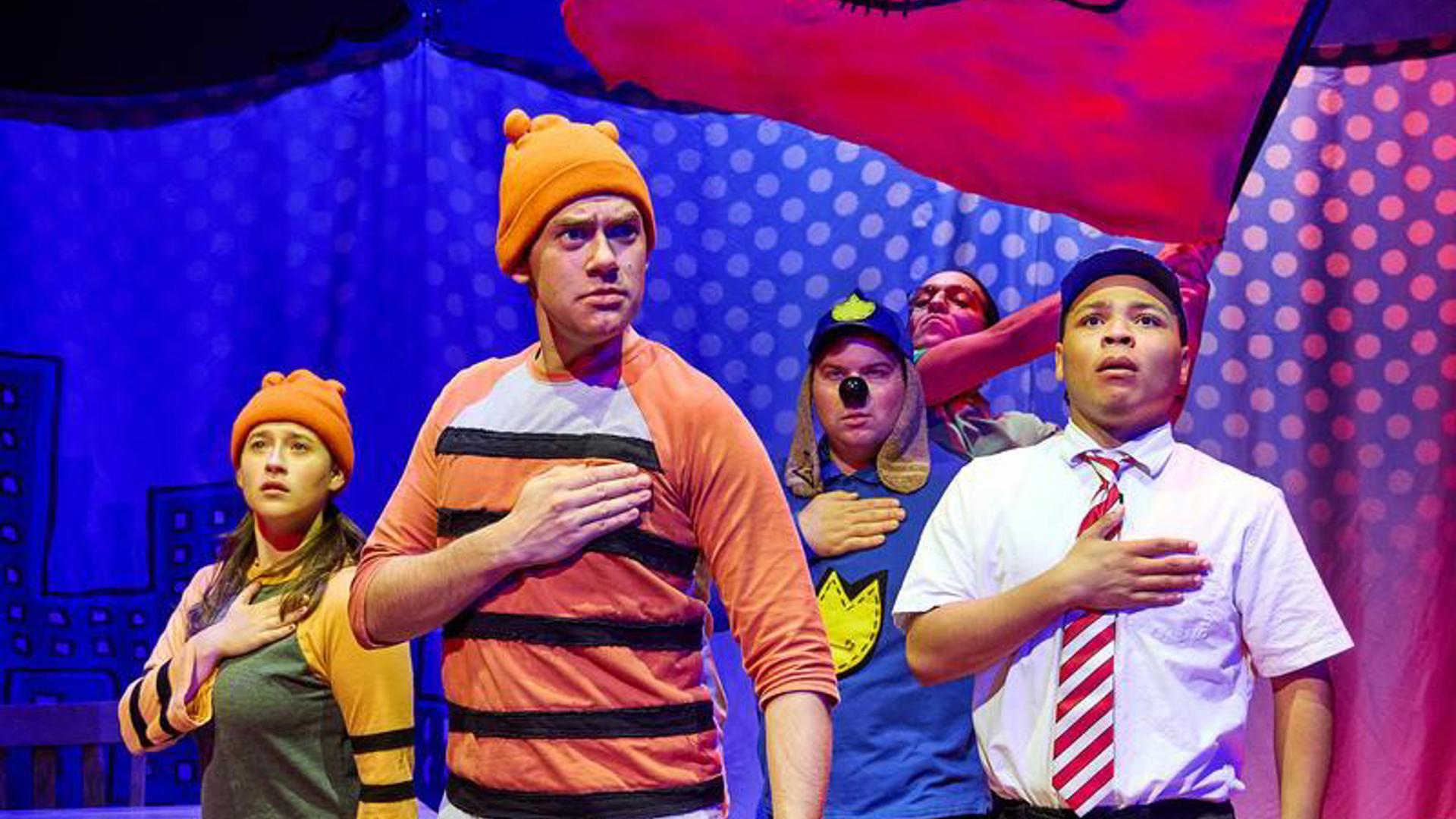 Five actors stand on stage, they all have their right hand on their hearts and the actor in the back is waving a flag with dog man's face on it.
