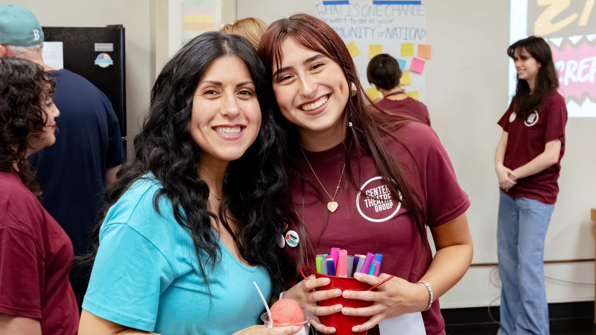 Two people pose, smiling, one is holding ice cream and the other is holding a small red bucket of markers. 