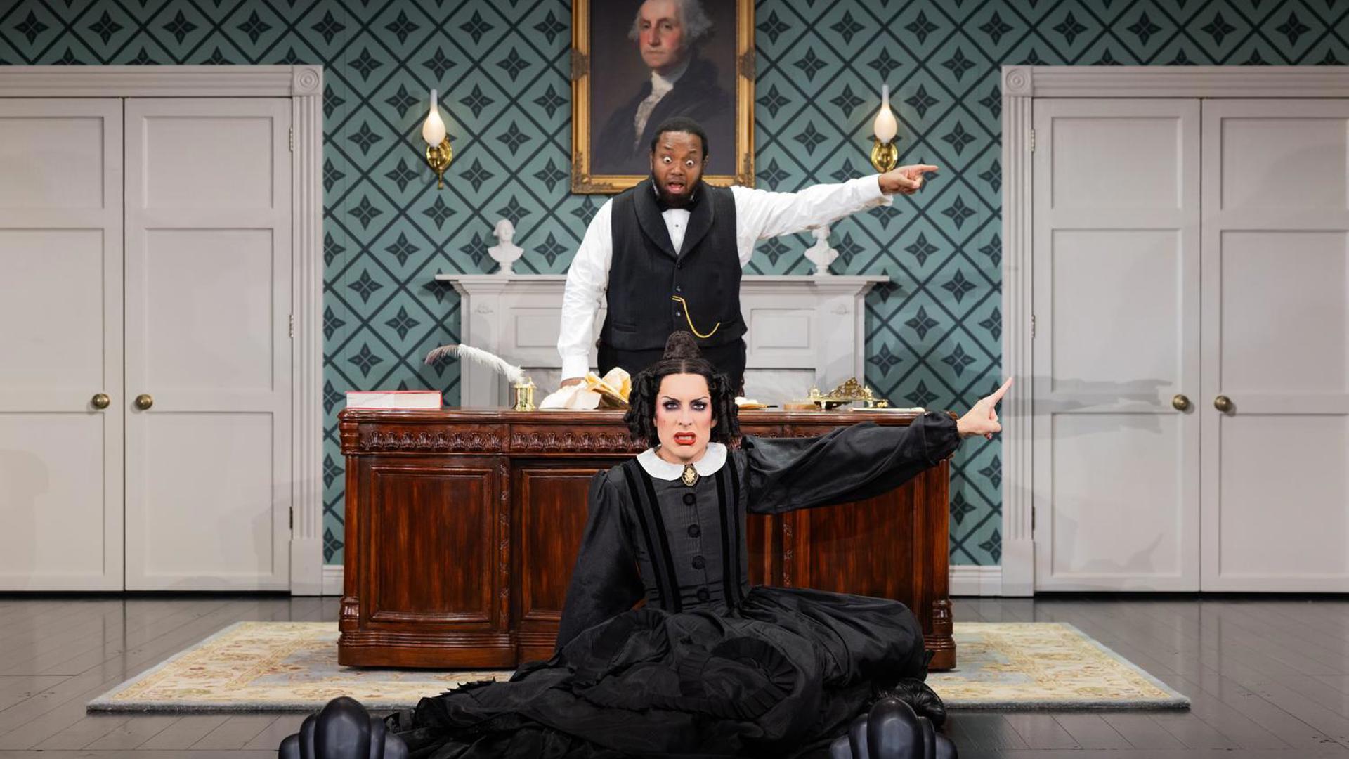 An actress in a black dress sits on a stage looking out at the audience, pointing to the left with her left hand. An actor stands behind her at a desk, looking at her and pointing the same way.