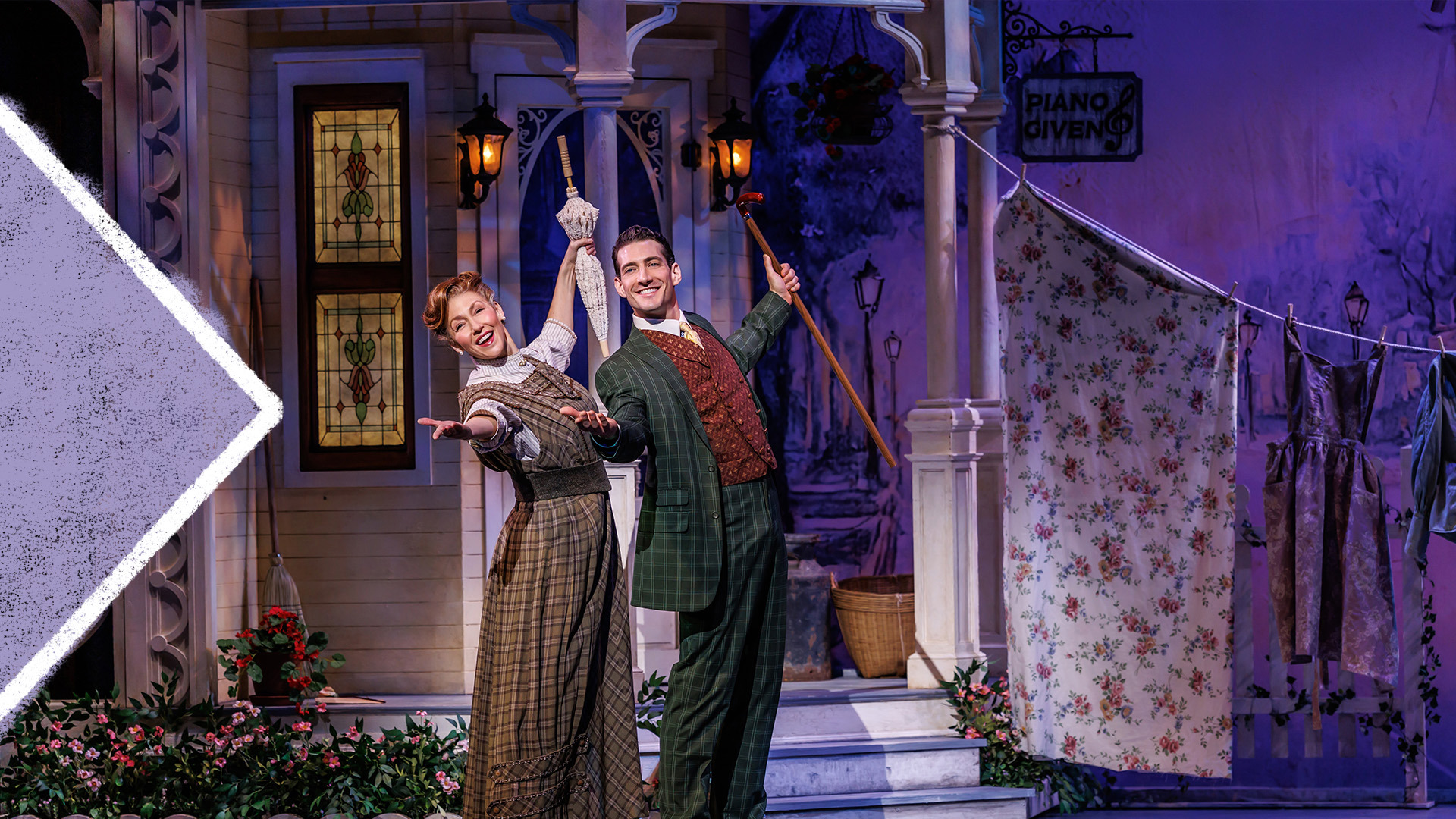 Two actors smiling post with their arts stretched out with one holding a closed umbrella and the other with a cane in front of wooden house.  