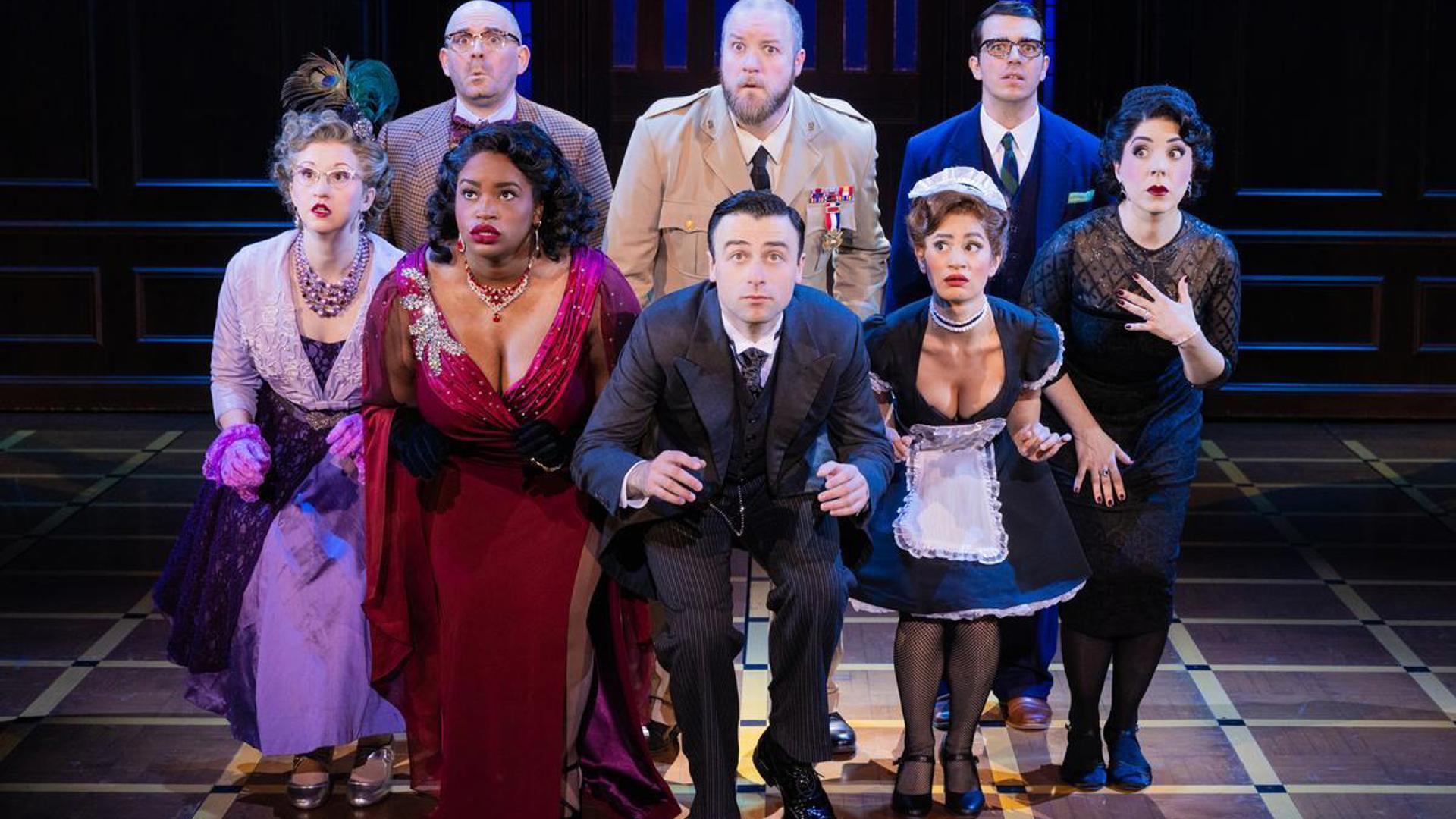 Four women and a man stand on stage crouched in a line with three men standing behind them as they search for something. All actors are looking to their forward off in the distance with a frightened look on their faces.
