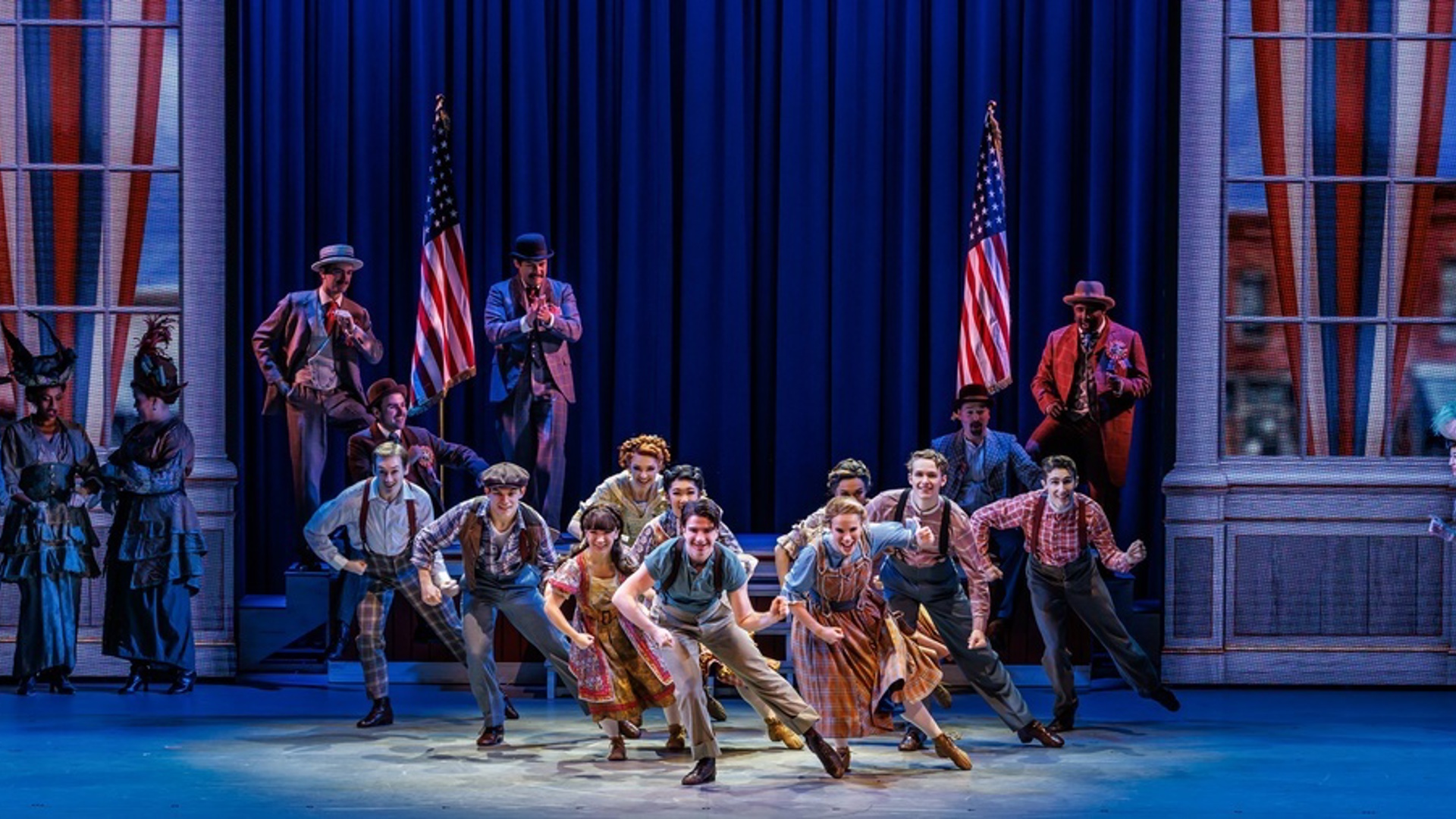 A group of actors dance in the middle of the stage in a V formation. There are two American flags behind them. There are actors watching them scattered on the right and left of the stage.